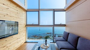 Een moderne woonkamer met een groot raam met uitzicht op zee, een flatscreen televisie aan de muur en een grijze bank.