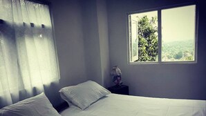 A bedroom with a bed, two pillows, a nightstand, a lamp, and a window with a view of greenery. - Mount view 11 (Kandy)