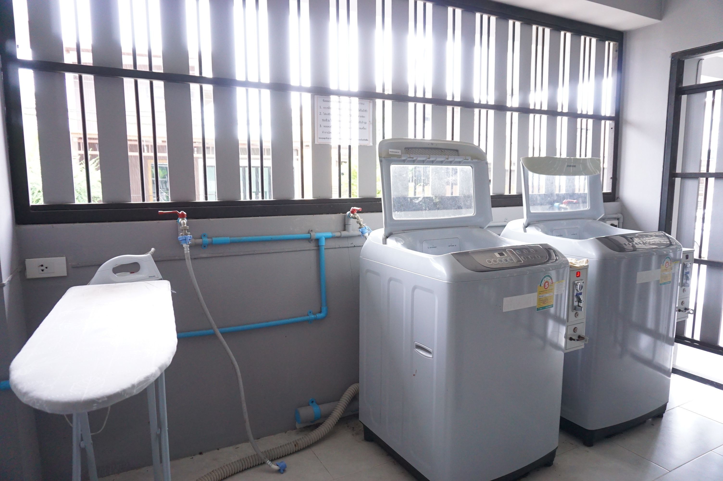 A laundry area with two large washing machines, a white ironing board, and a window with vertical blinds.