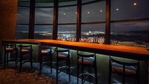 A bar area with a long counter and multiple bar stools, large windows offering a view of a cityscape at night.