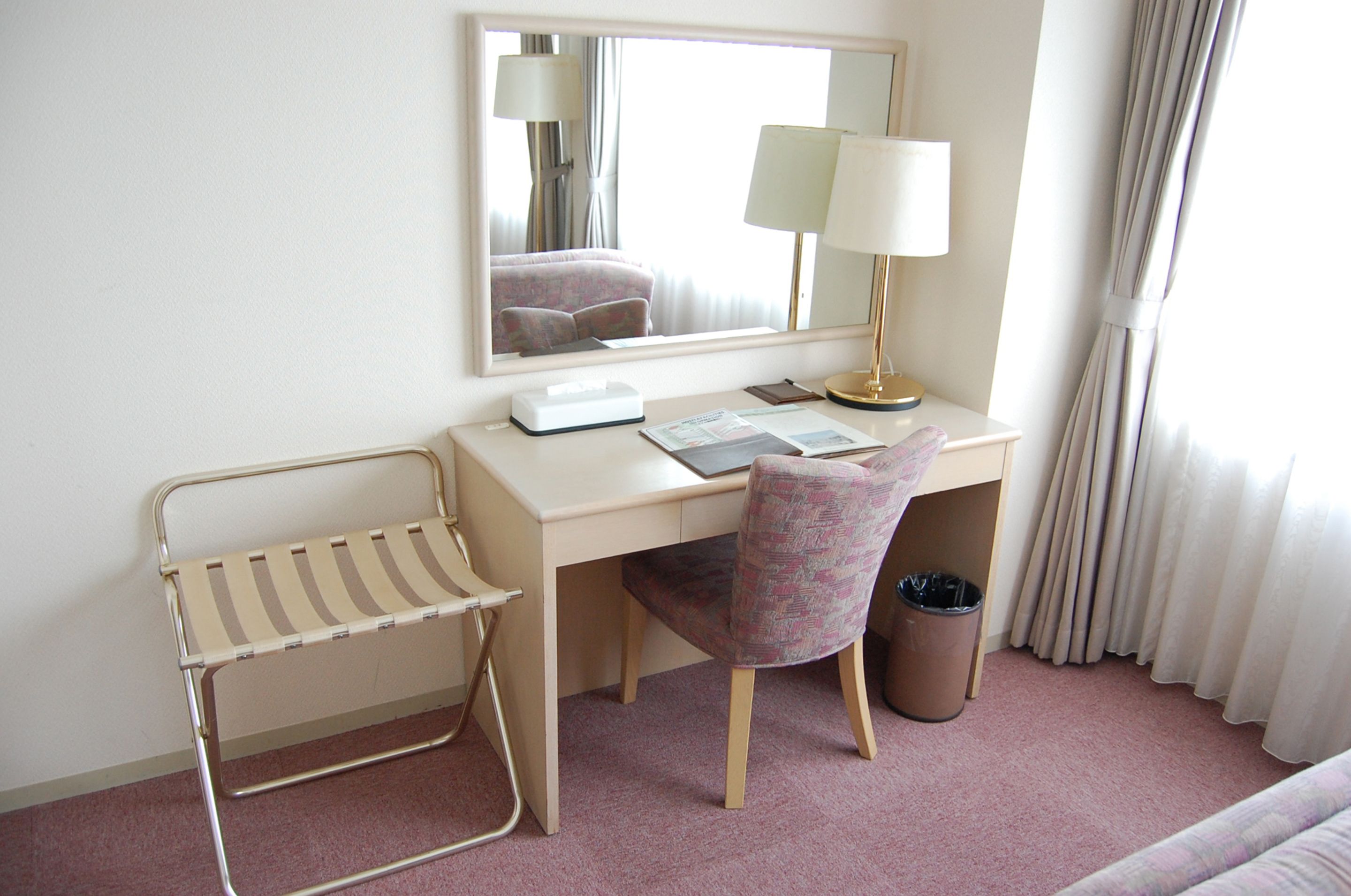 Habitación de hotel con escritorio, silla, lámpara, espejo y ventana con cortinas.
