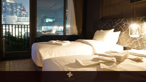 Een hotelkamer met een groot bed, een nachtkastje met lamp en uitzicht op een stadsbeeld door een groot raam.