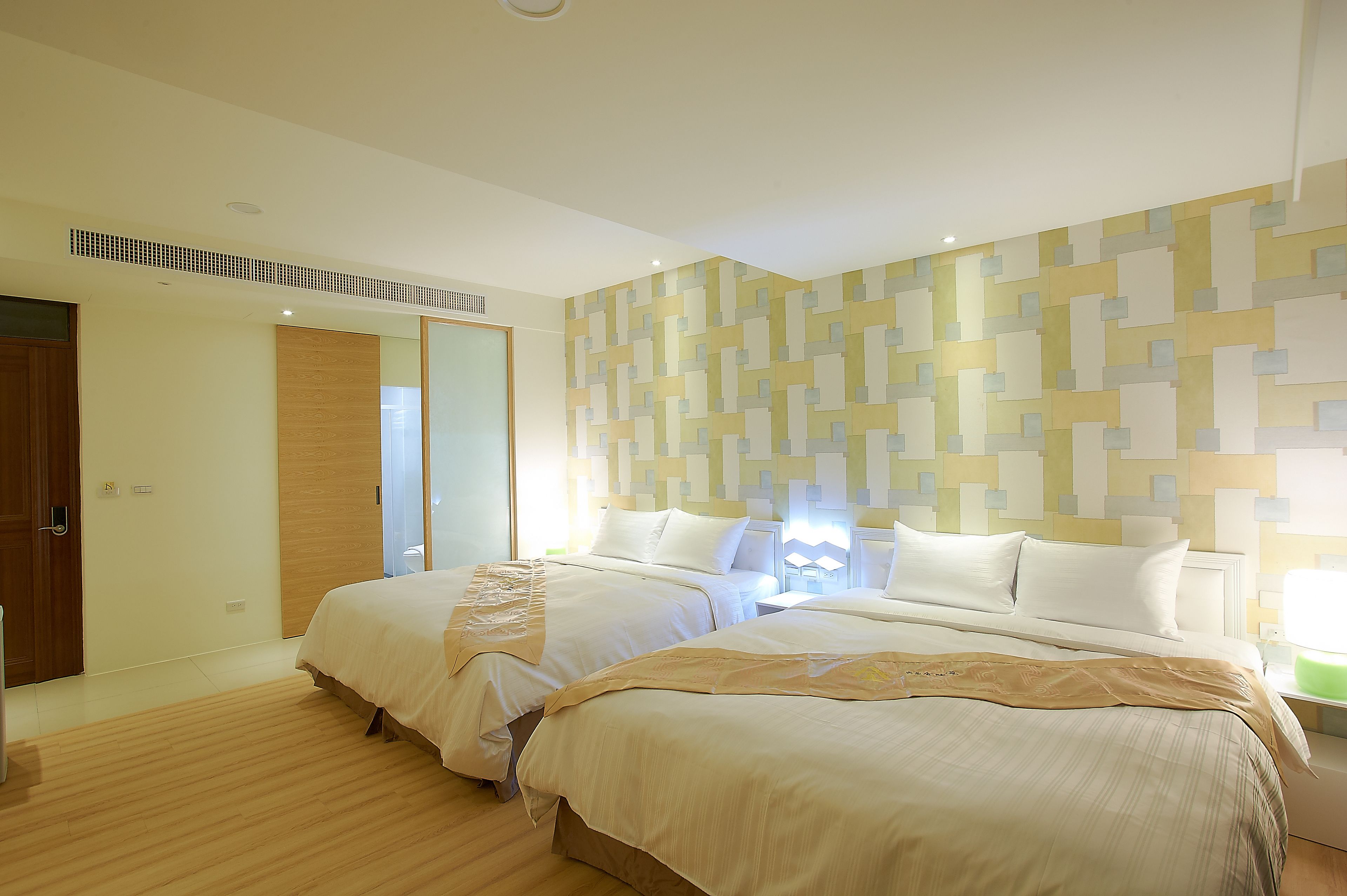 A hotel room with two beds, a wooden floor, a patterned wall, and a door.