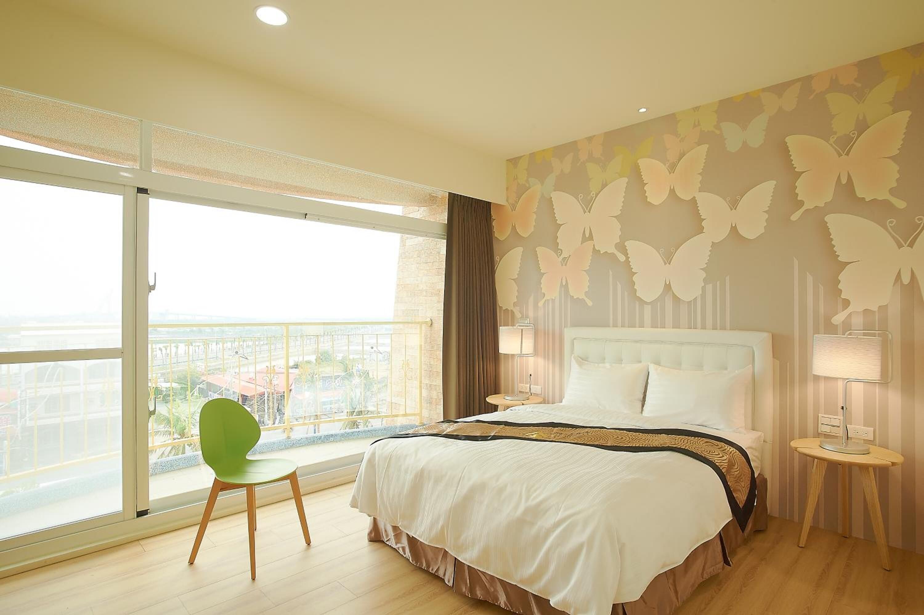A hotel room with a large bed, a green chair, a balcony with a view, and butterfly wall art.