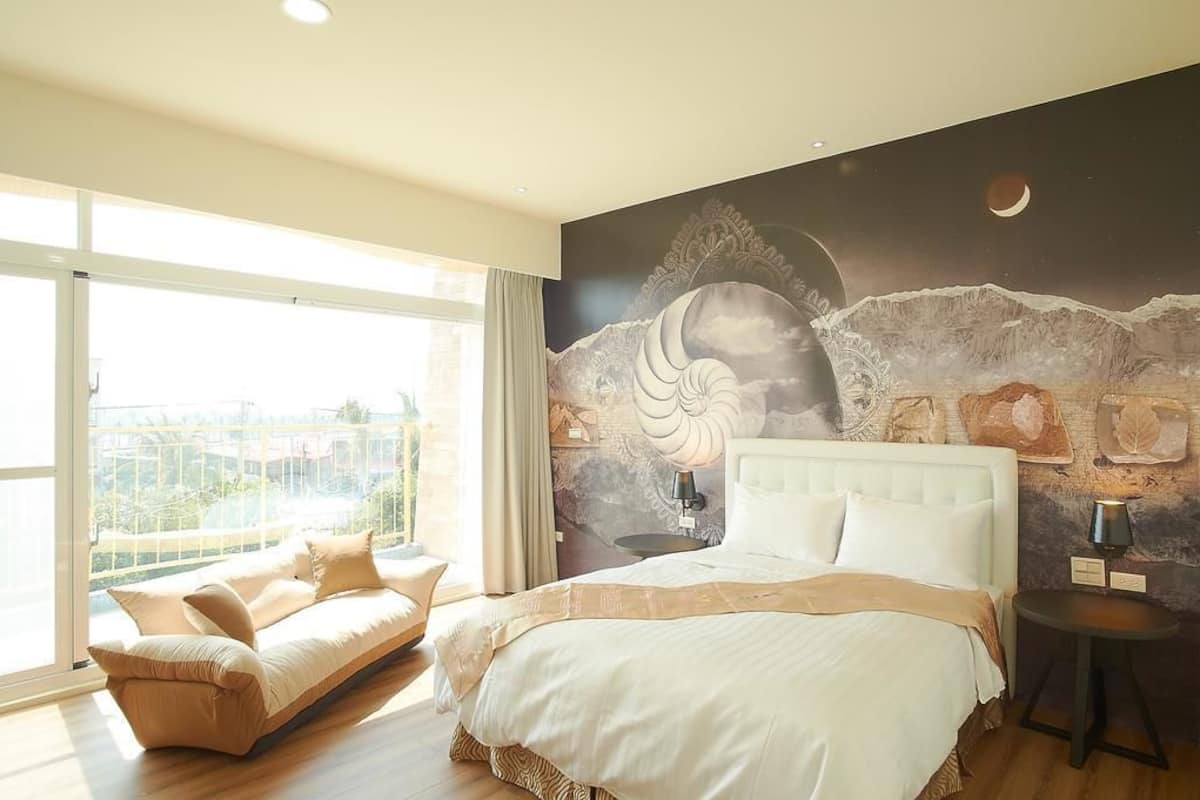A bedroom with a large bed, a sofa, a view through a balcony, and a mural on the wall.