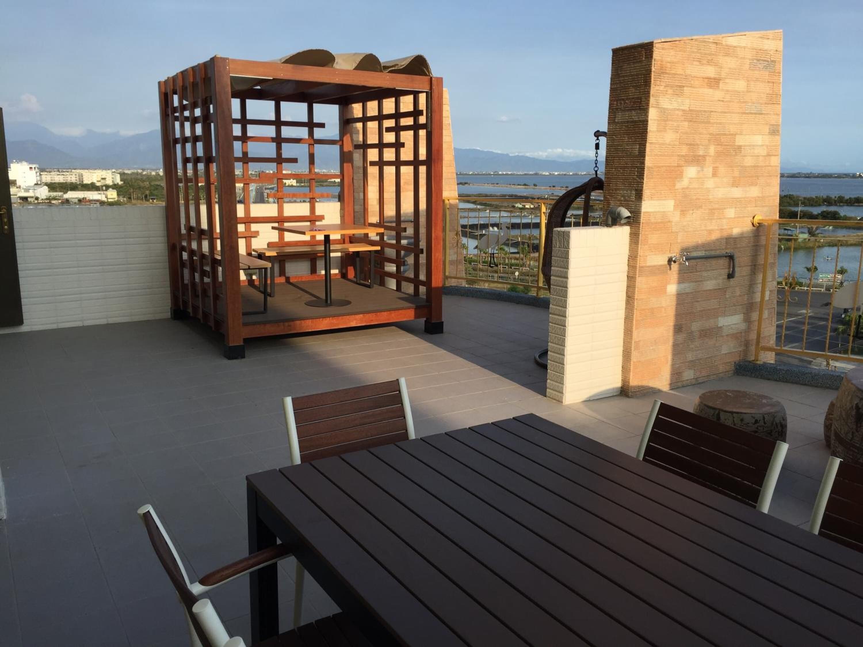 A rooftop terrace with a wooden table and chairs, a wooden partition with seating, and a brick wall with a window overlooking a body of water and distant mountains.