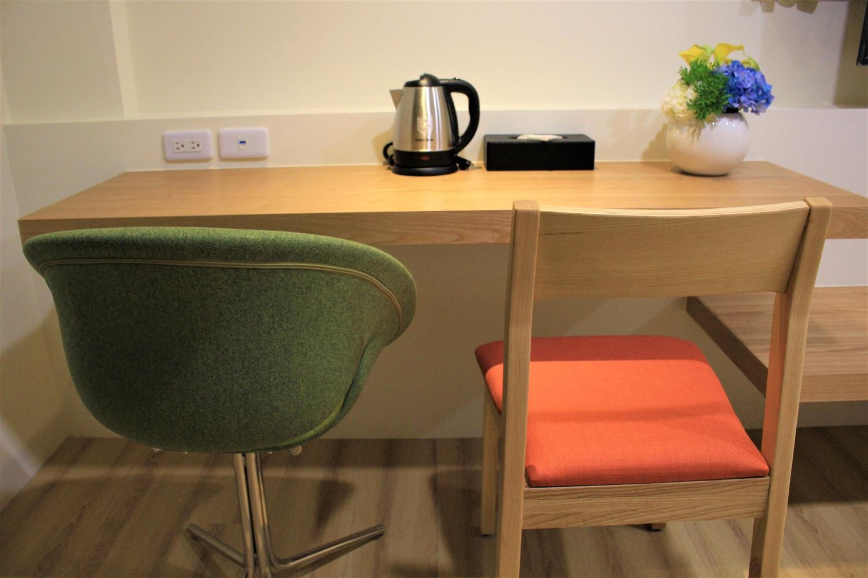 A wooden table with a kettle, a vase with flowers, and a chair with a green backrest and orange seat cushion.