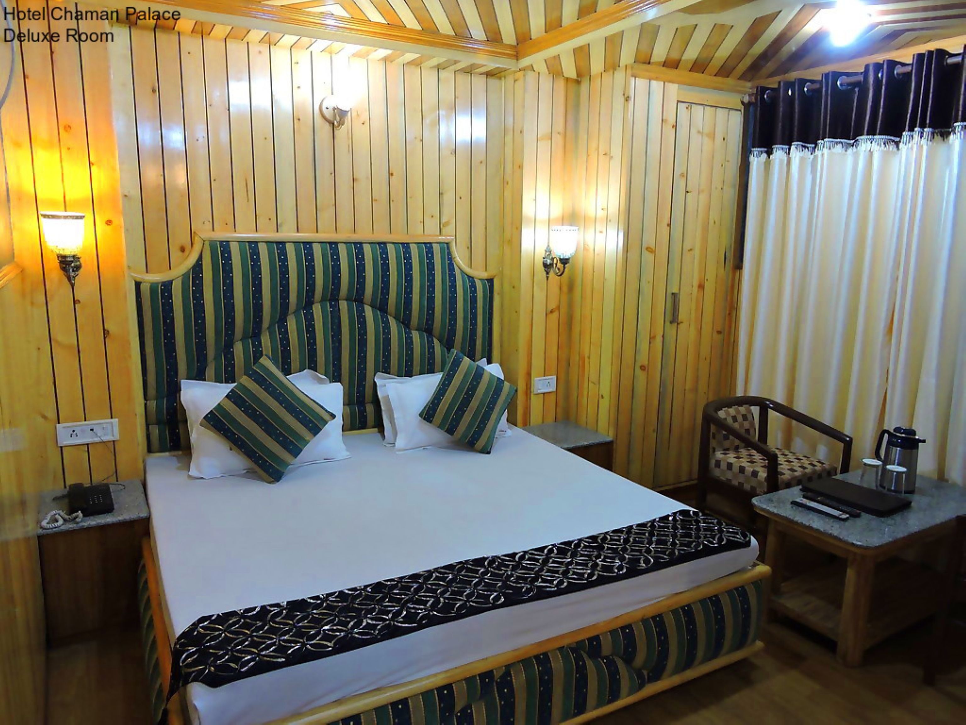A Deluxe Room in Hotel Chaman Palace with a wooden paneled wall, a bed with patterned bedding, bedside lamps, and a small table with a kettle.
