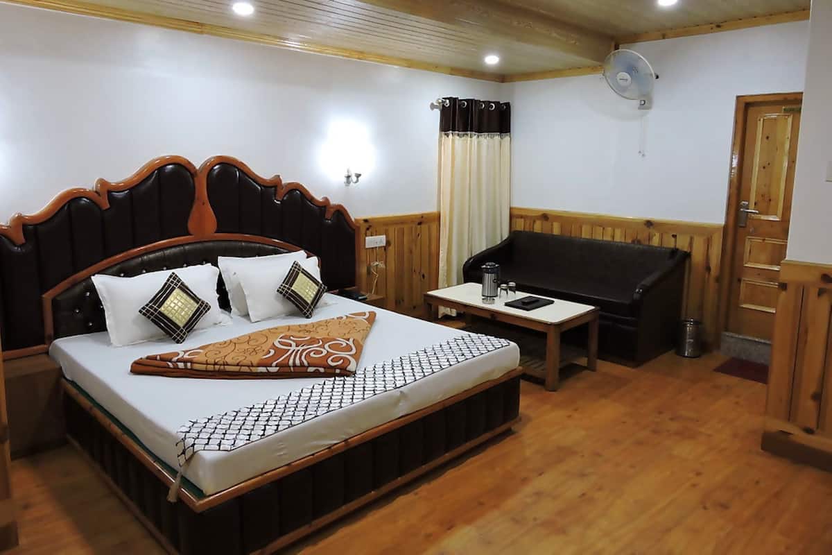 A hotel room with a large bed, a wooden headboard, a small table with a kettle, a sofa, and a fan.