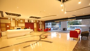 A modern hotel lobby with a reception desk, red and grey seating, and a large mirror reflecting the space. - Hotel Jump In & Out (Coimbatore)