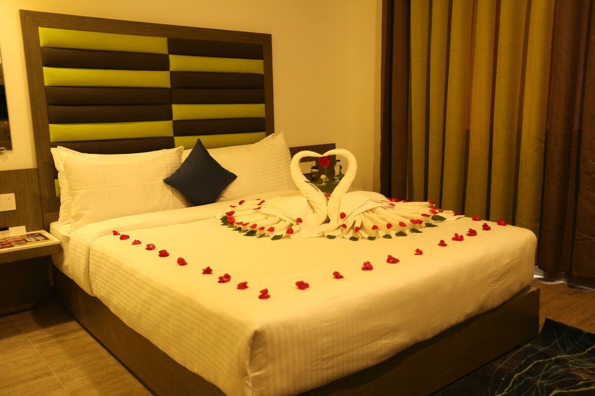 A neatly made bed with white linens, a heart-shaped towel arrangement, and rose petals on the bed.