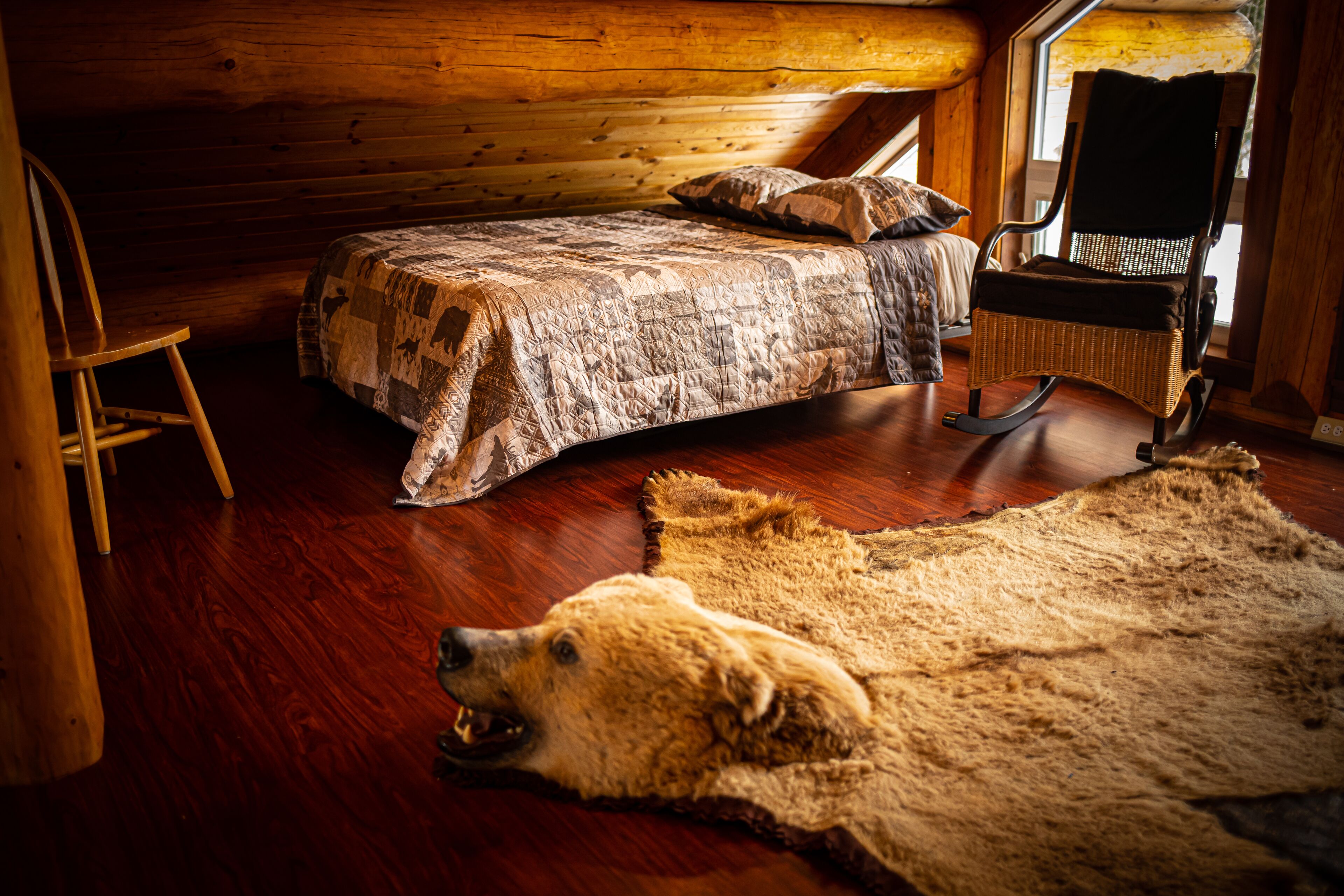 A cozy cabin interior with a wooden bed, a rocking chair, and a large fur rug.