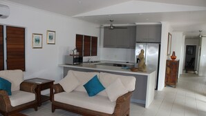A modern kitchen with grey cabinets, a central island, and stainless steel appliances. There is a wicker sofa with blue cushions in the living area. - Banfields Retreat (Wongaling Beach)