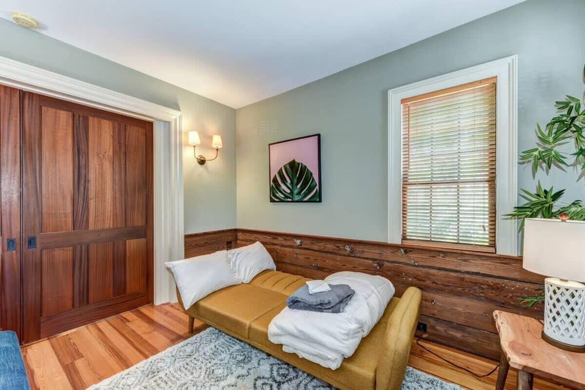 A bedroom with a wooden headboard, a bed with white and beige bedding, a wooden wardrobe, a window with blinds, and a wall-mounted lamp.