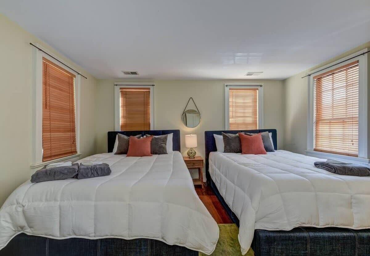 A room with two beds, each with white bedding and different colored pillows. There are wooden blinds on the windows, and a small table with a lamp between the beds.