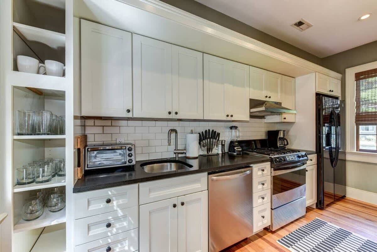 A modern kitchen with white cabinets, a black countertop, stainless steel appliances, and a window with blinds.