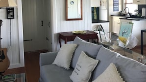 Een gezellige woonkamer met een grijze bank, een houten salontafel en een lijstvormige spiegel aan de muur. Er is een keuken te zien op de achtergrond, met witte keukenkastjes en een raam.