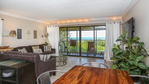Een woonkamer met een houten tafel, een bank en een televisie. Er is een balkon met uitzicht op de oceaan.