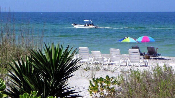 Een strand met ligbedden en parasols, een boot op het water, en heldere luchten.