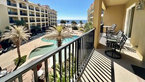 A balcony with a view of a pool and beach, surrounded by palm trees and a hotel building.