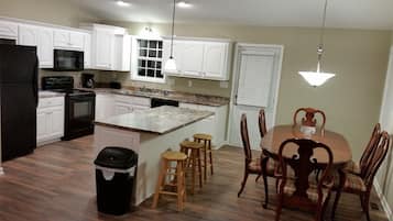 A kitchen with white cabinets, a black refrigerator, a microwave, and a stove. There is a wooden dining table with four chairs and a trash can.