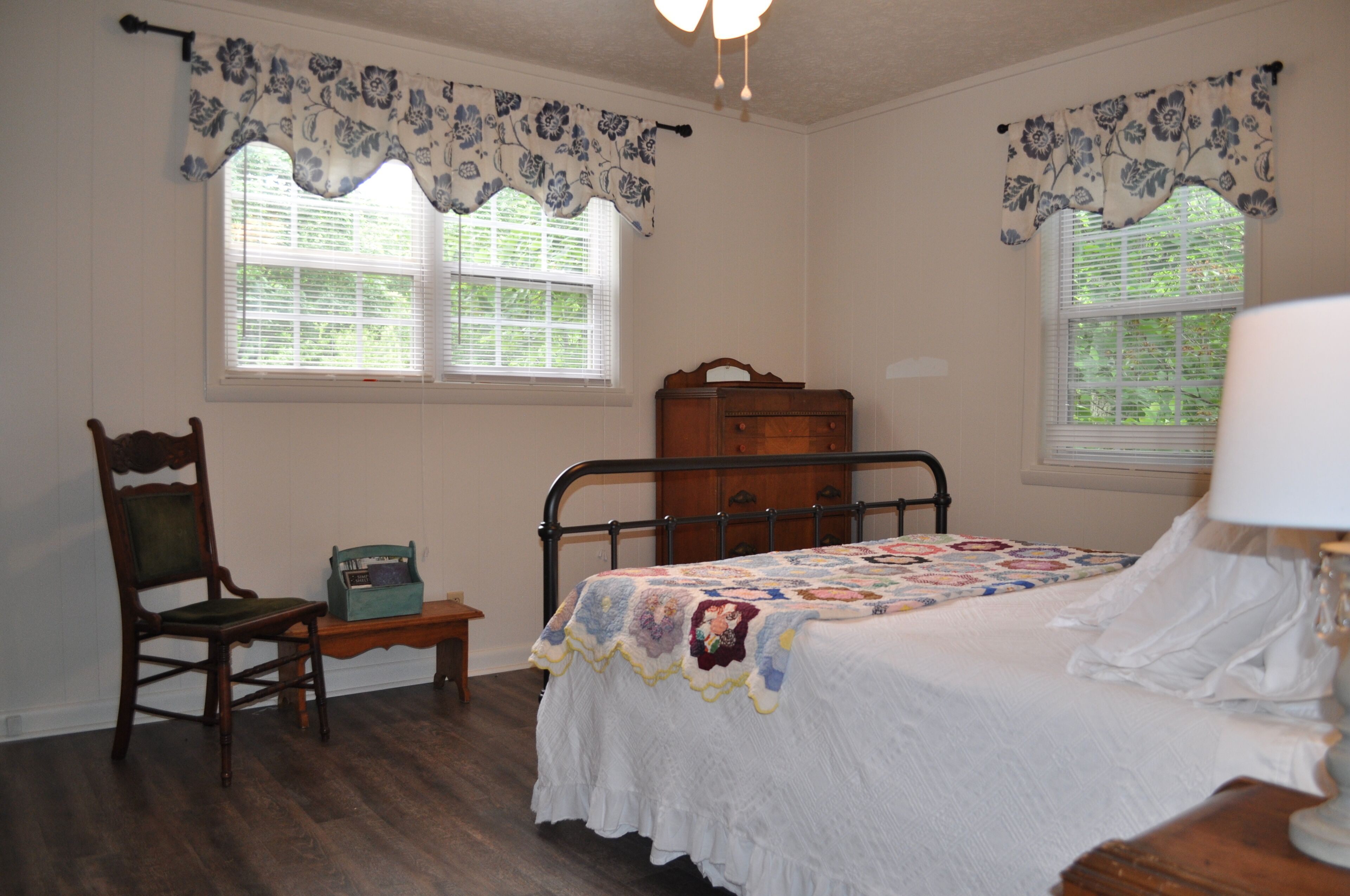 A bedroom with a bed, a chair, a dresser, and two windows with floral curtains.