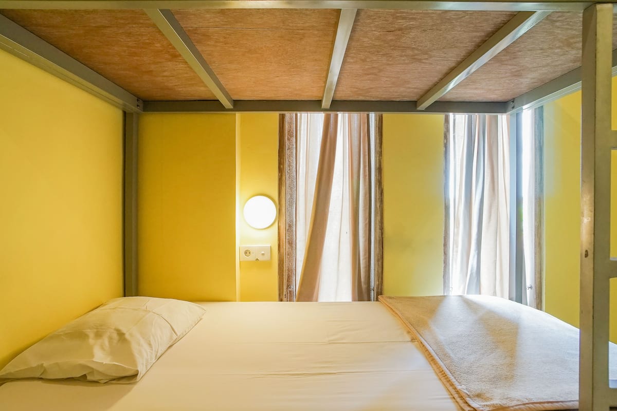 A room with yellow walls, a wooden ceiling, a bed with white bedding, a round wall clock, and a window with curtains.