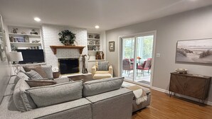 A living room with a grey sectional sofa, a fireplace, a bookshelf, and a dining area with chairs and a table. - 'Stones Throw' Lakeside Home - Walk to Marina + Town + Assoc Beach (New Buffalo)