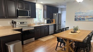 A modern kitchen with dark cabinets, stainless steel appliances, and a wooden dining table with chairs. - 'Stones Throw' Lakeside Home - Walk to Marina + Town + Assoc Beach (New Buffalo)