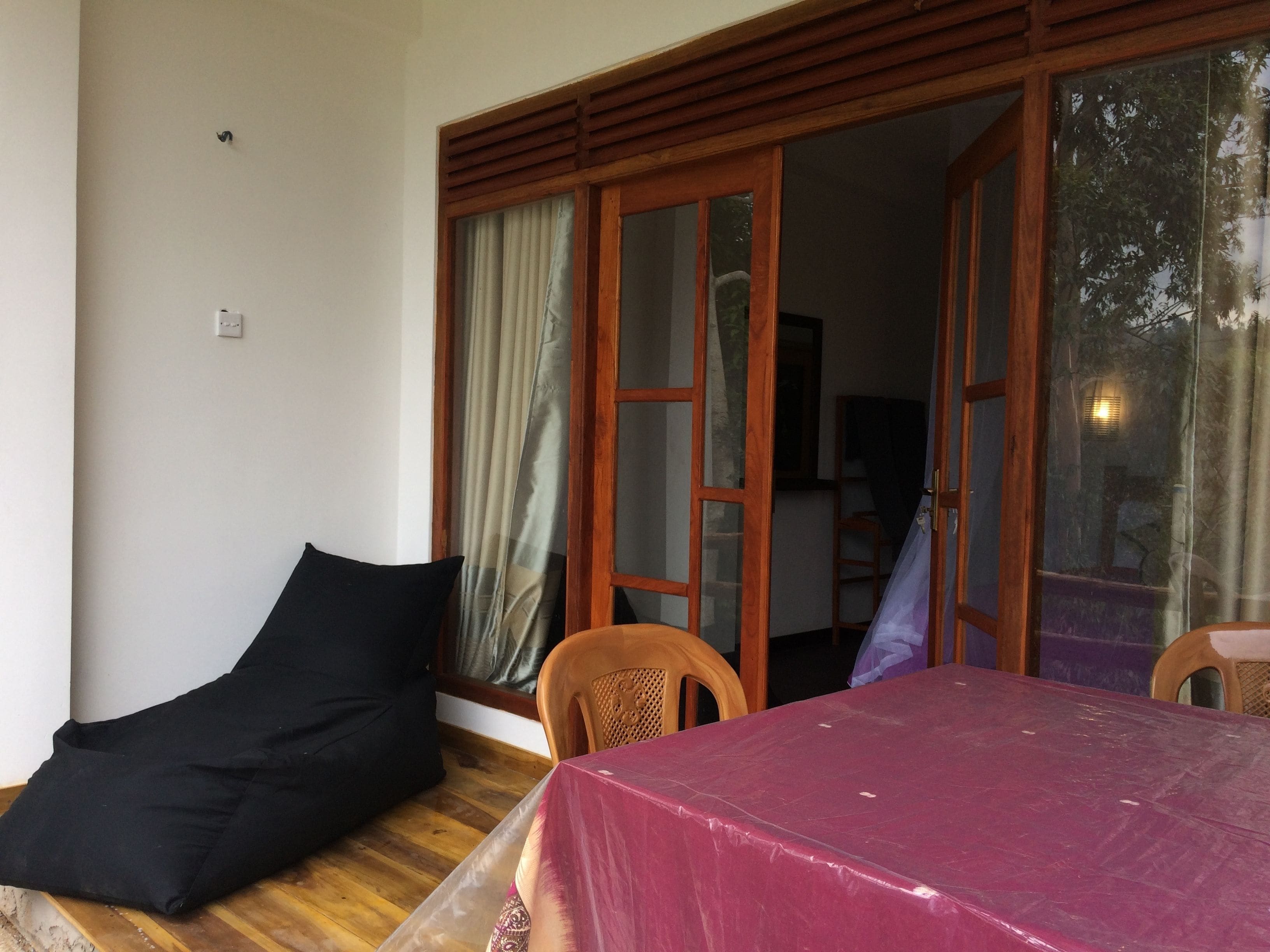 A room with a wooden floor, a black cushion, a table with a red tablecloth, and a sliding glass door leading to an outdoor area.