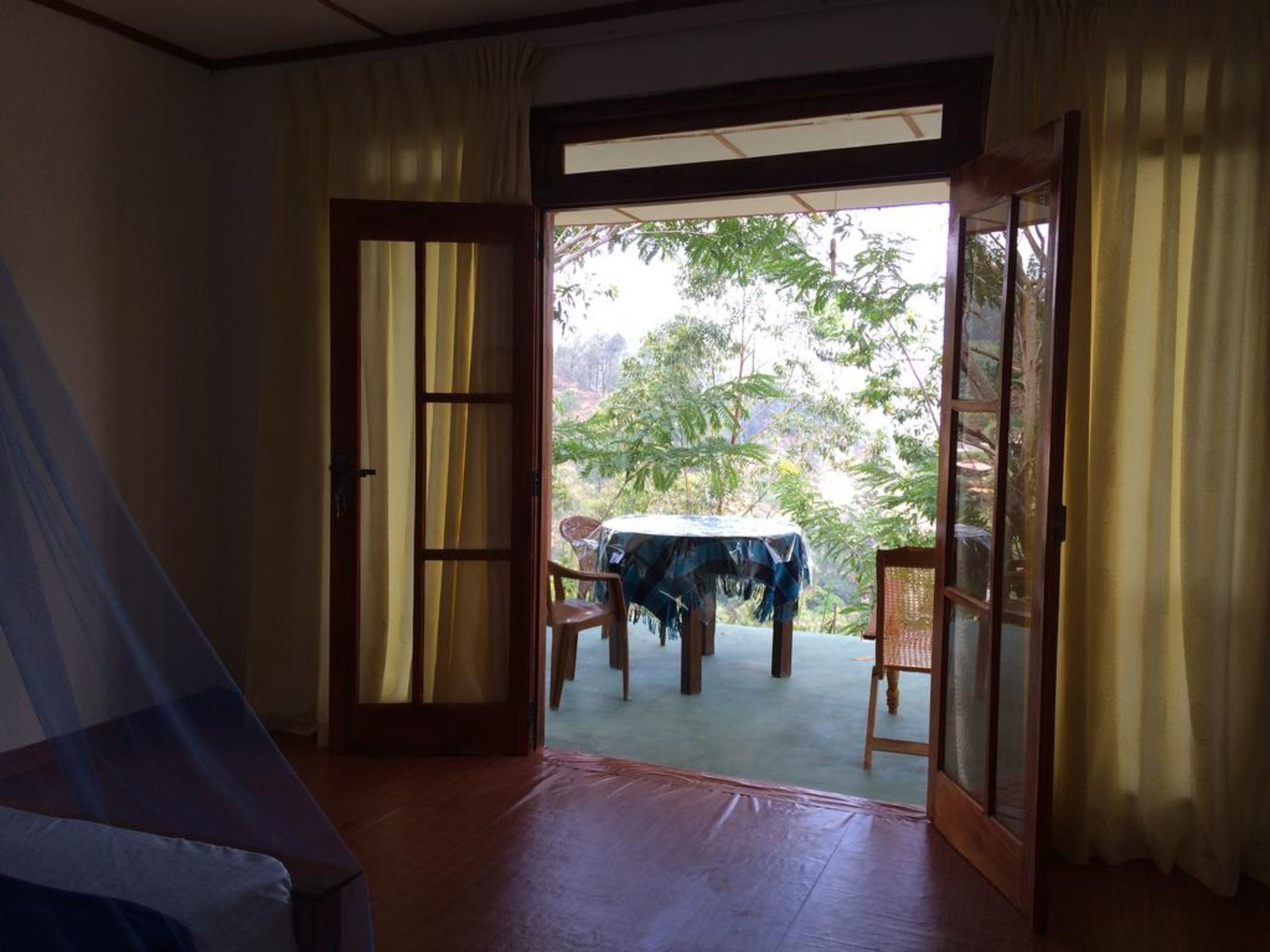 A room with a wooden floor, a bed, a table with a tablecloth, and a view of a garden through an open door.