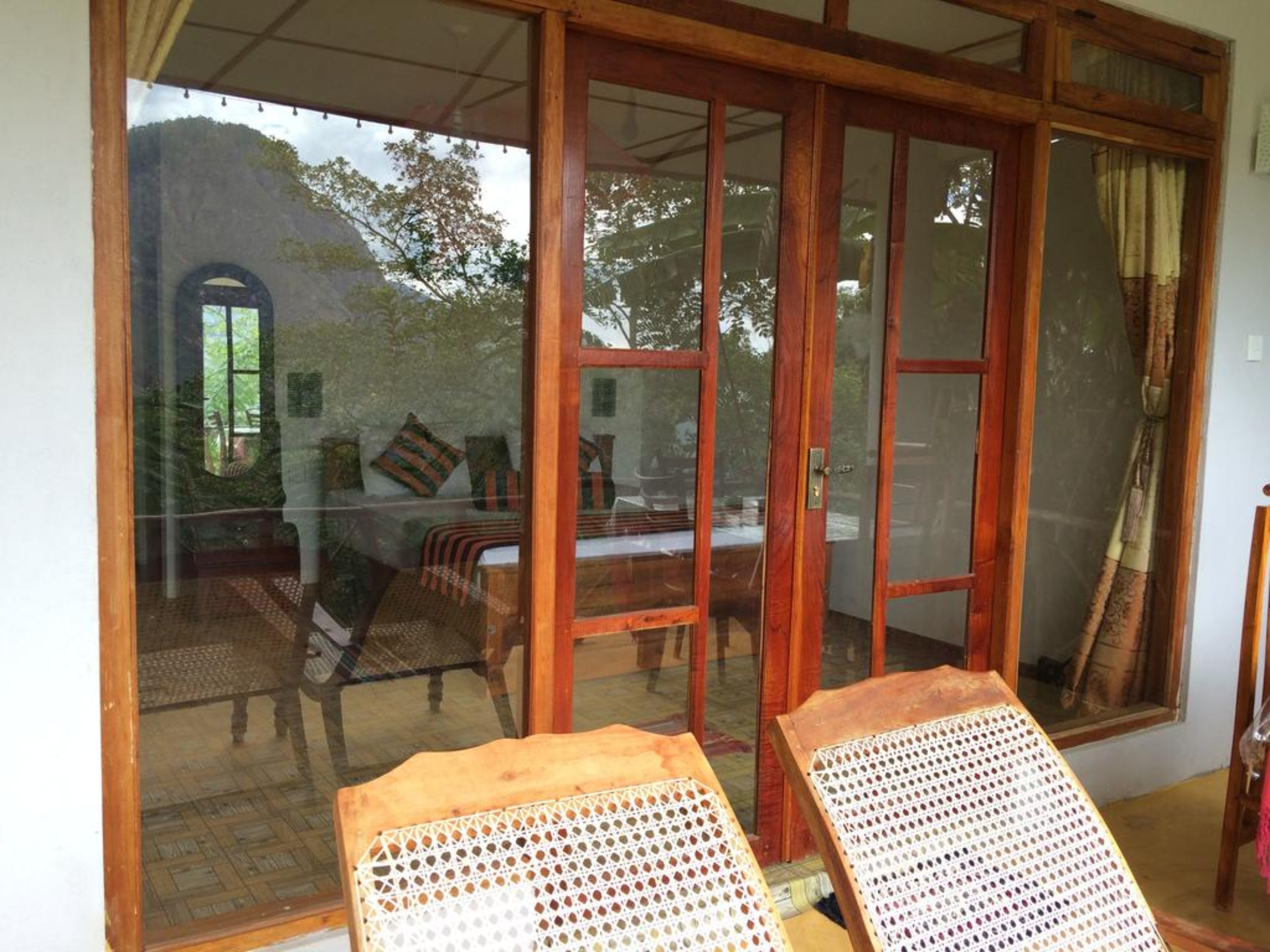 A room with a large sliding glass door leading to an outdoor area with a view of trees and a building. Inside, there are two wicker chairs and a table.