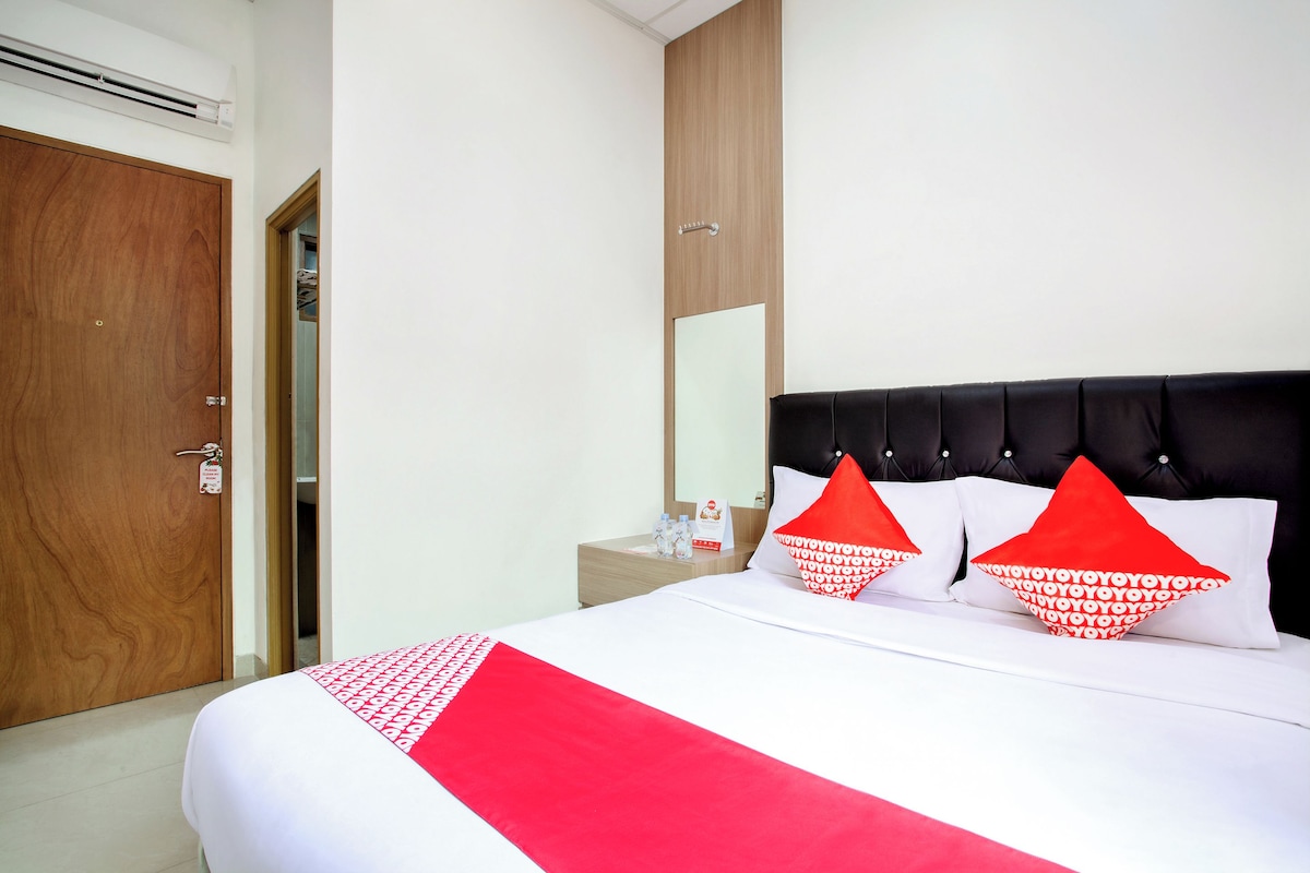 Habitación de hotel con una cama grande, una puerta de madera, una mesita de noche con espejo y una colcha roja y blanca.