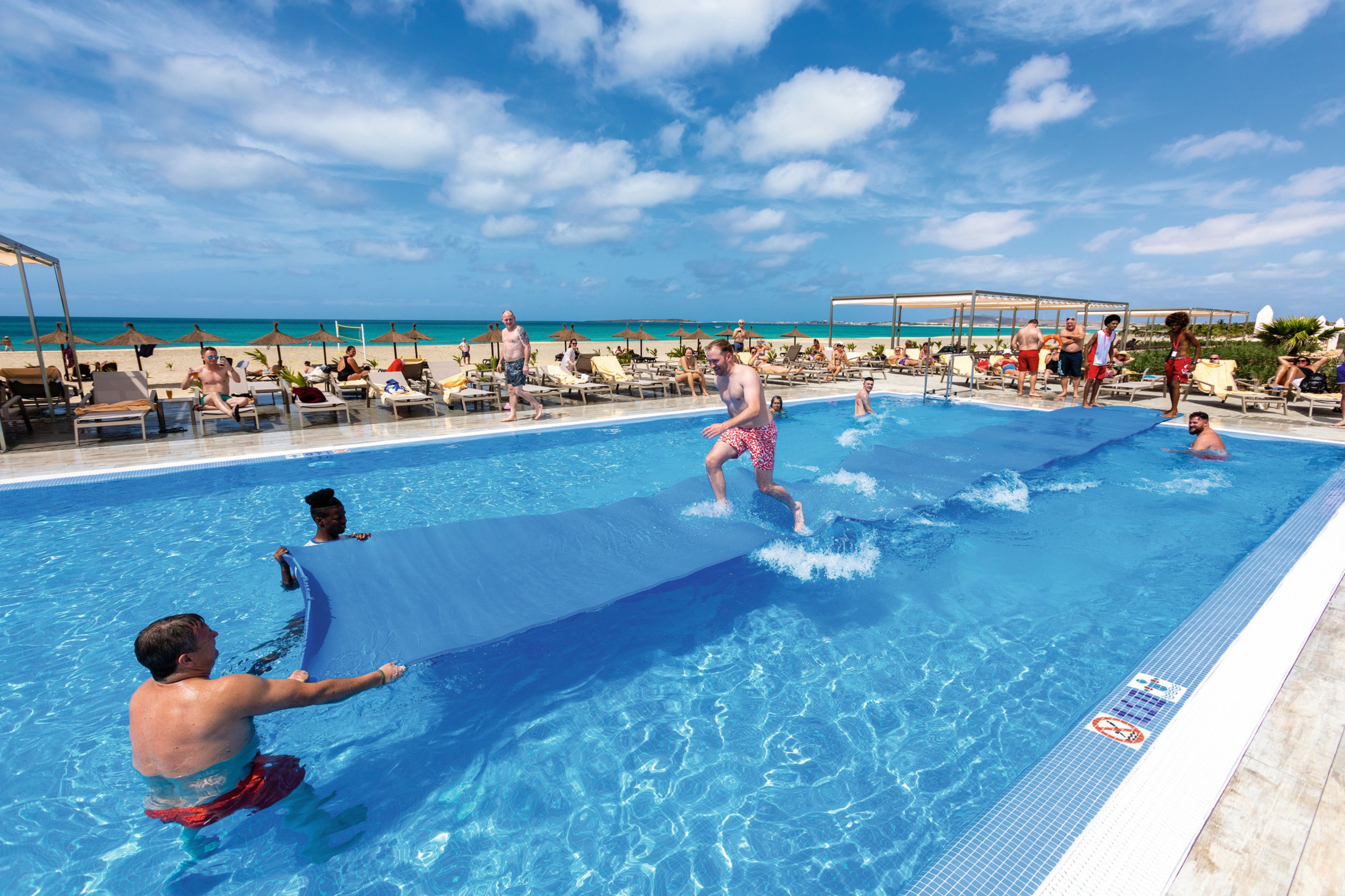 A pool area with lounge chairs and umbrellas, a clear sky, and a beach in the background.