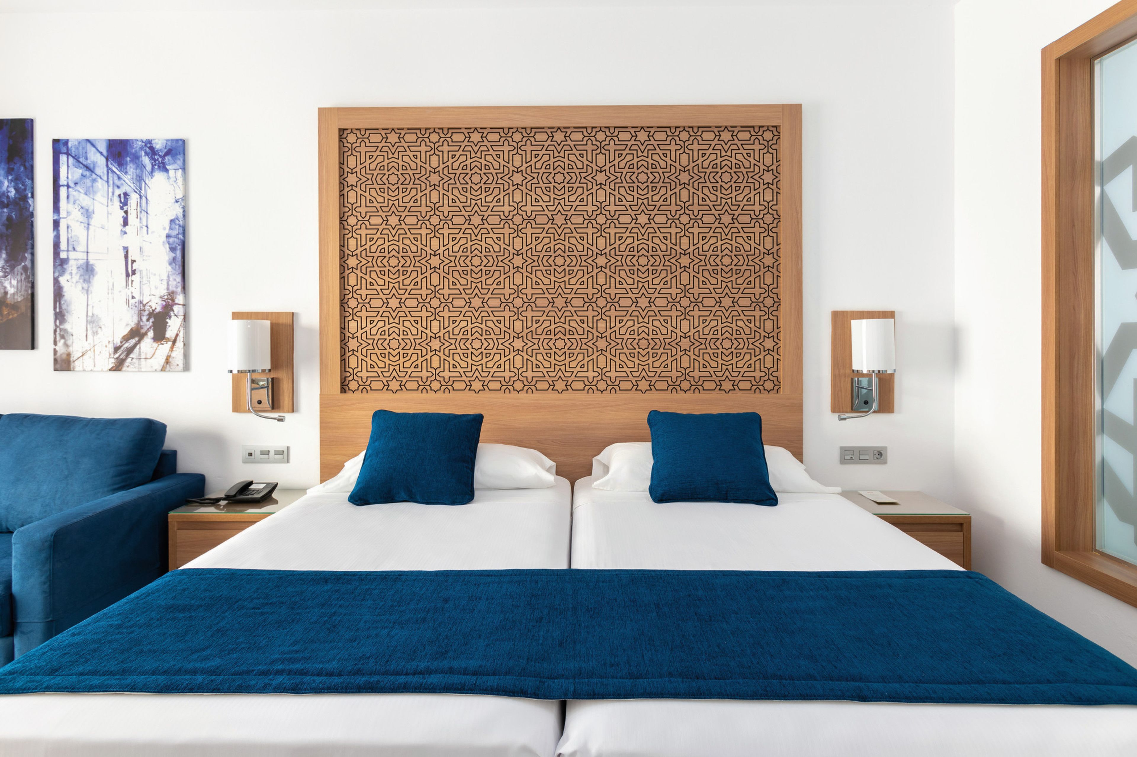 A hotel room with a large bed, two blue pillows, a wooden headboard with a geometric pattern, two bedside tables with lamps, and a blue chair.