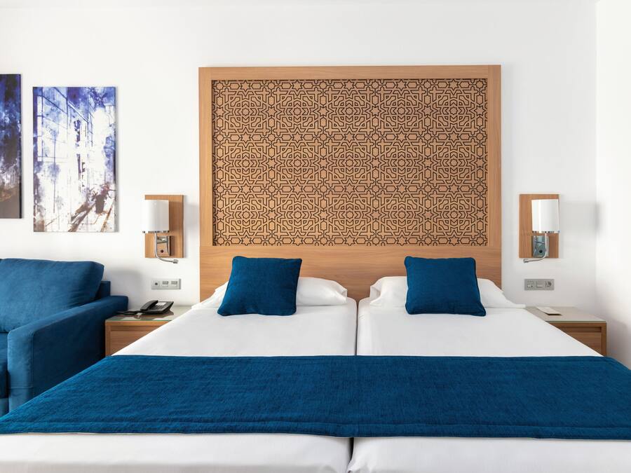 A hotel room with a large bed, two blue pillows, a wooden headboard with a geometric pattern, two bedside tables with lamps, and a blue chair.