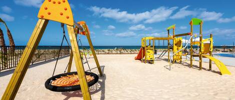 Une aire de jeux sur la plage avec des balançoires et des toboggans, une zone délimitée par une clôture et une plage de sable.