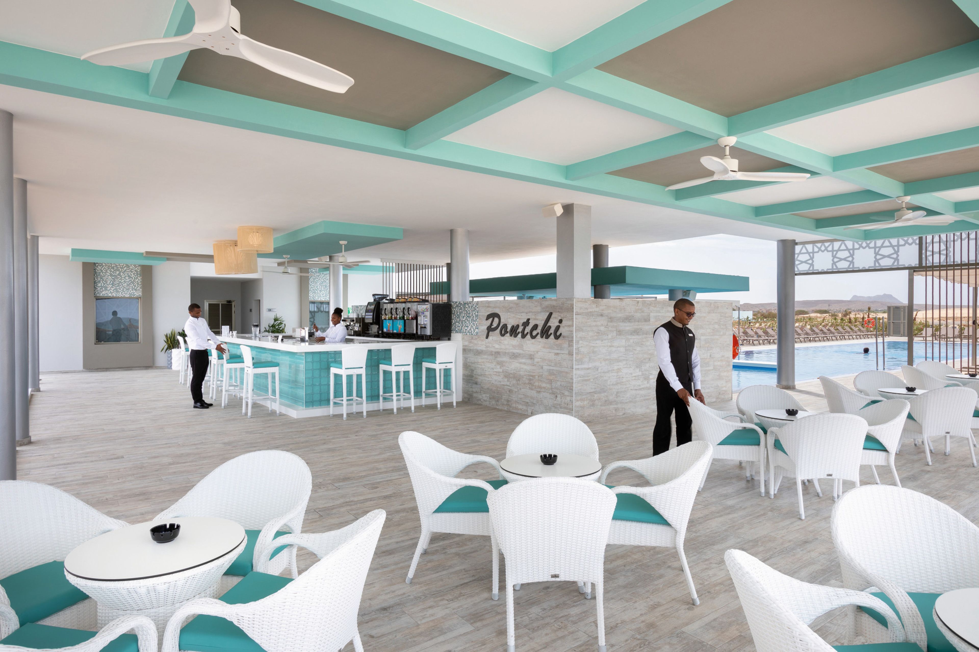 A modern outdoor seating area with white wicker furniture and teal cushions, a bar area with a bartender, and a view of the landscape.