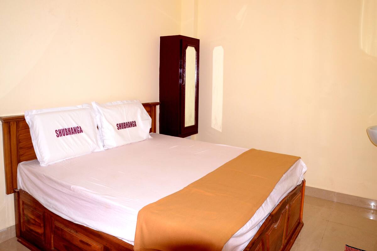 A neatly made bed with white bedding and two pillows labeled "SHUBHANGA".