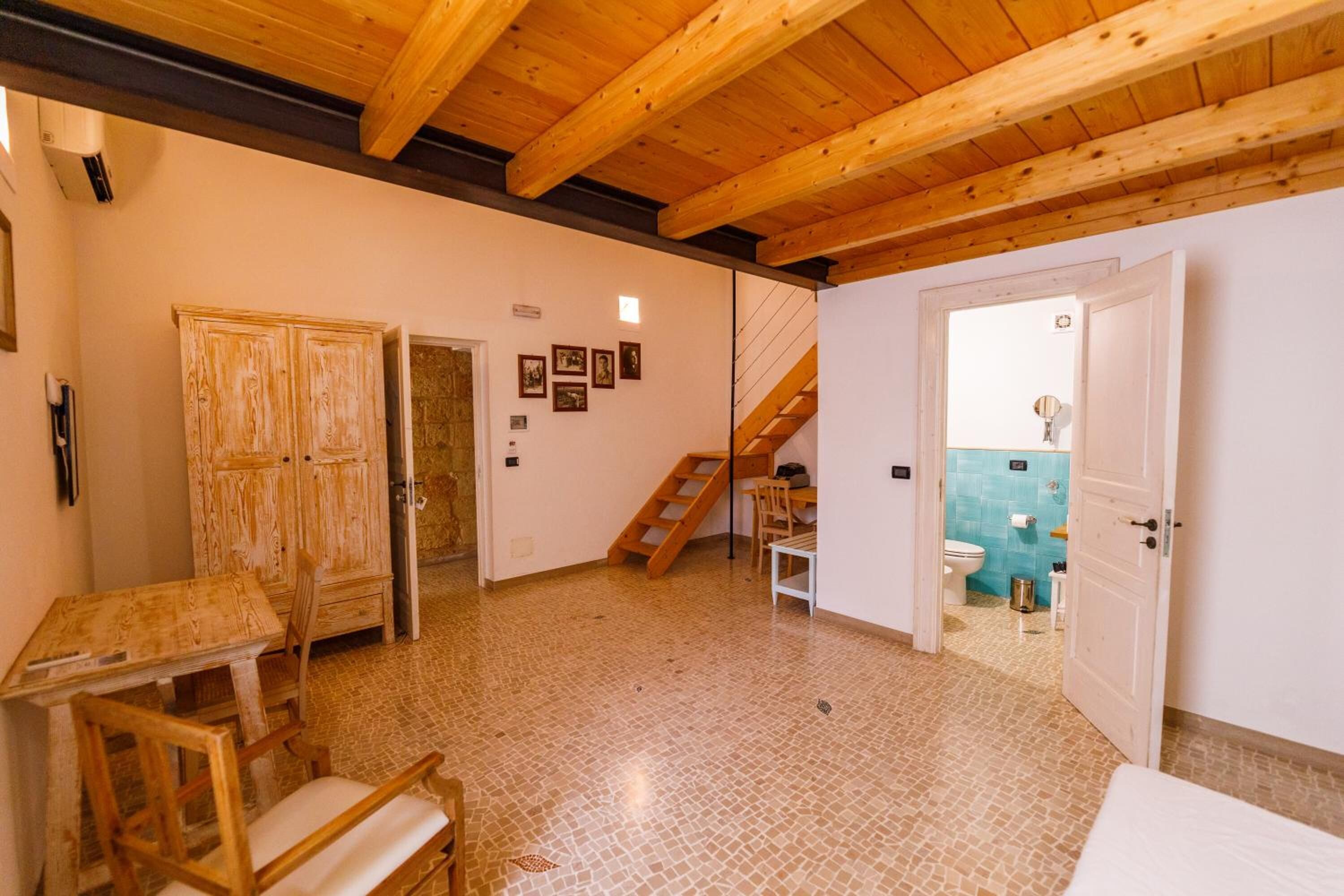 A room with a wooden ceiling, a tiled floor, a wooden table with chairs, a wardrobe, a staircase, and a bathroom visible through an open door.