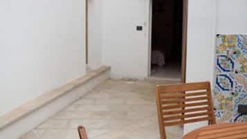 Une terrasse avec une table ronde en bois et des chaises, un sol carrelé et un mur blanc avec une porte donnant sur une pièce intérieure.