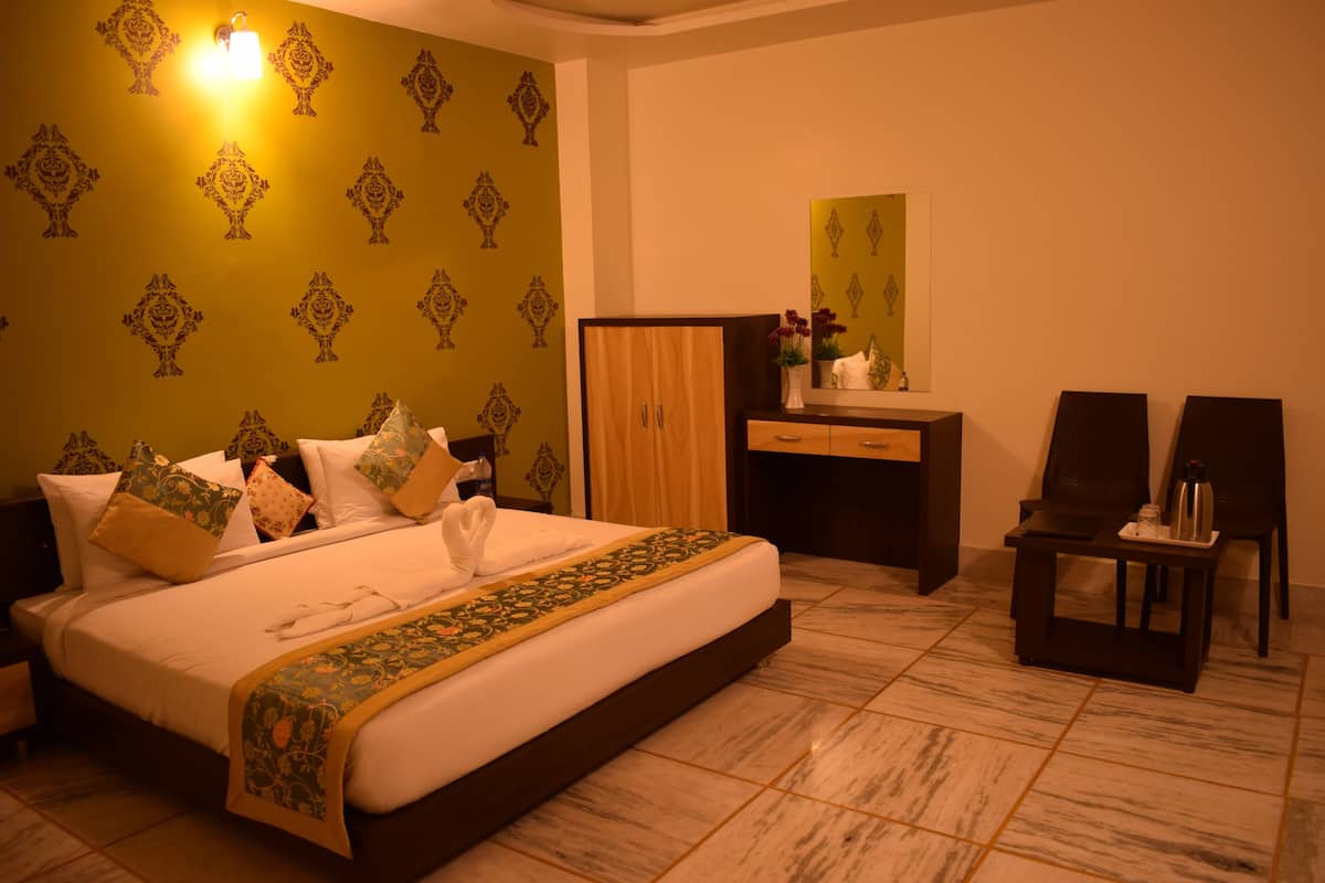 A hotel room with a large bed, a wooden wardrobe, a desk with a chair, and a small table with a bottle.