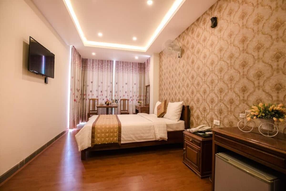 A hotel room with a bed, bedside table, a small desk with a telephone, a television mounted on the wall, and a patterned wallpaper.