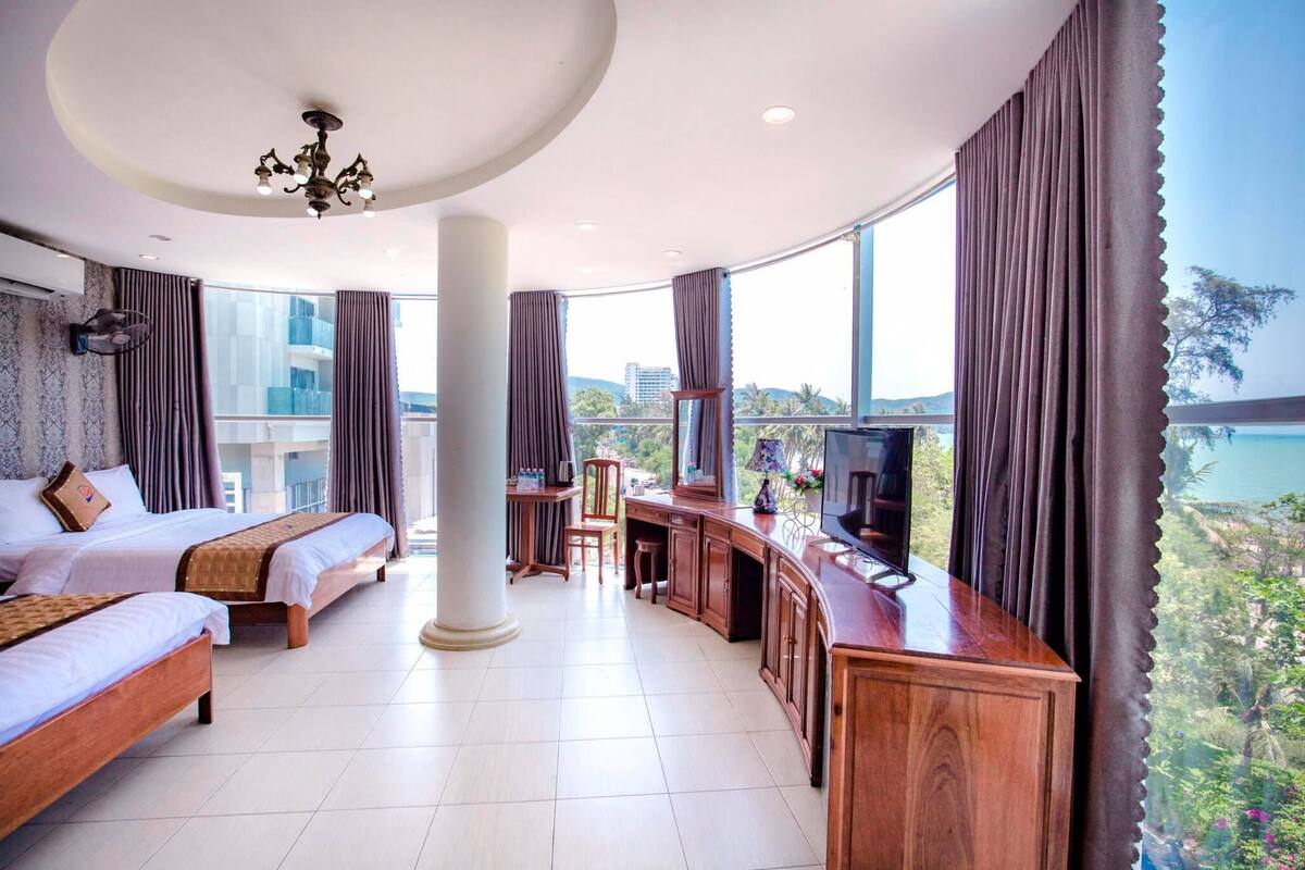A hotel room with two beds, a TV, a desk, and a view of the sea.