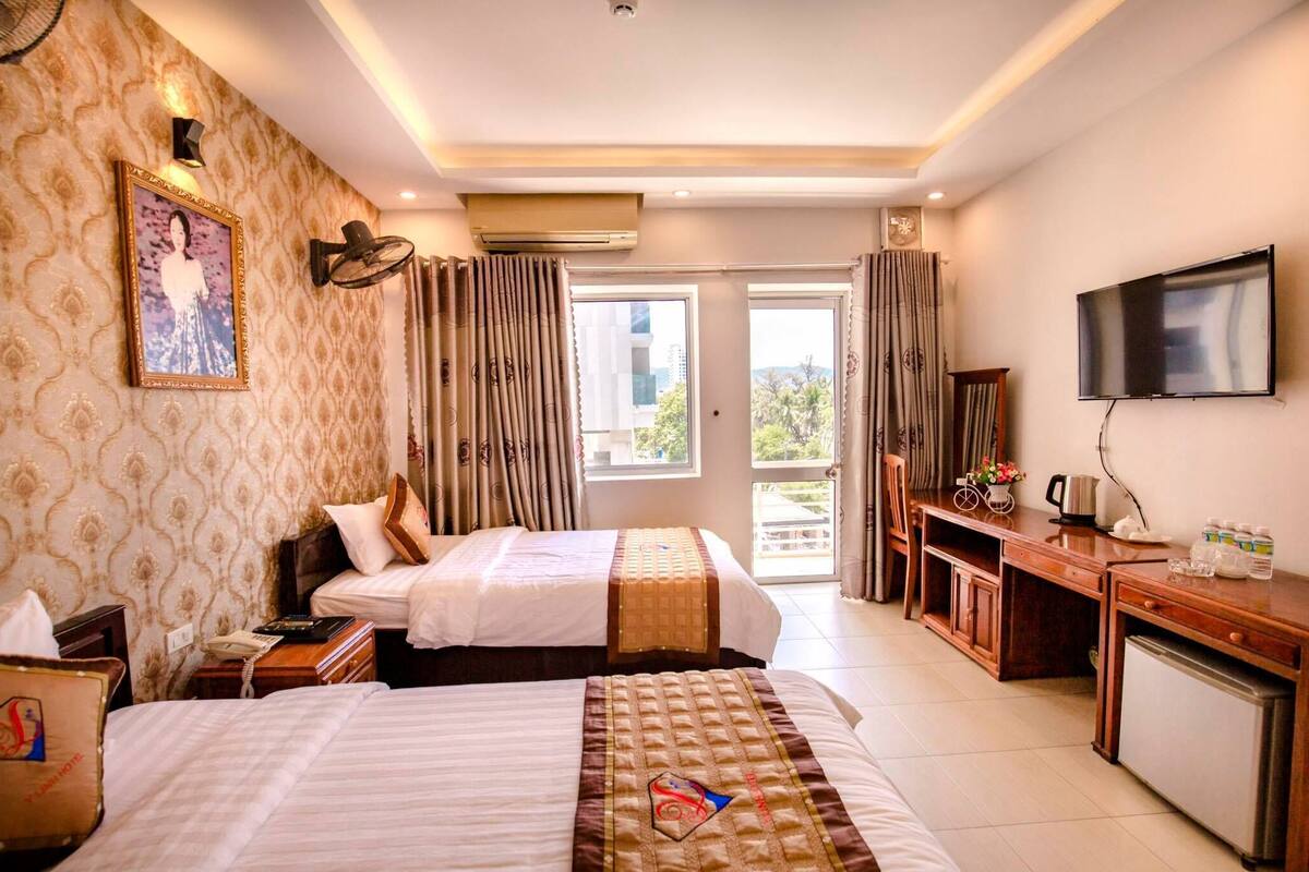 A hotel room with two beds, a desk with a TV, a small fridge, and a window with curtains.