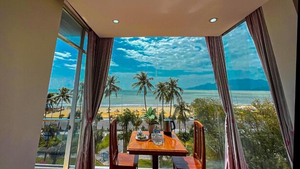A room with a large window overlooking a beach, a table with chairs, and a view of palm trees and the ocean. - Y Linh Hotel (Quy Nhon)