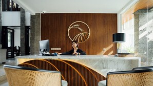 A reception desk with a woman seated behind it, a computer, and a decorative wall piece. There are two wicker chairs in front of the desk. - Umpadhi Canggu Hotel (Canggu)