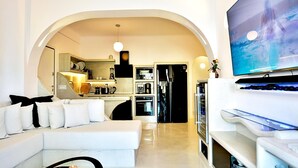 Una sala de estar moderna con un sofá blanco, un televisor de pantalla plana y una zona de cocina con refrigerador y horno.