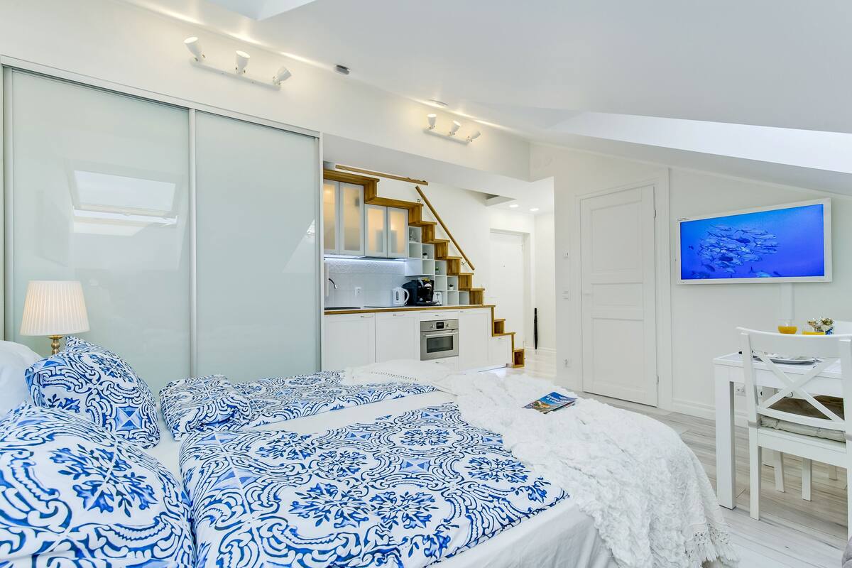 A modern bedroom with a large bed, a small kitchenette, a staircase, and a TV on the wall.