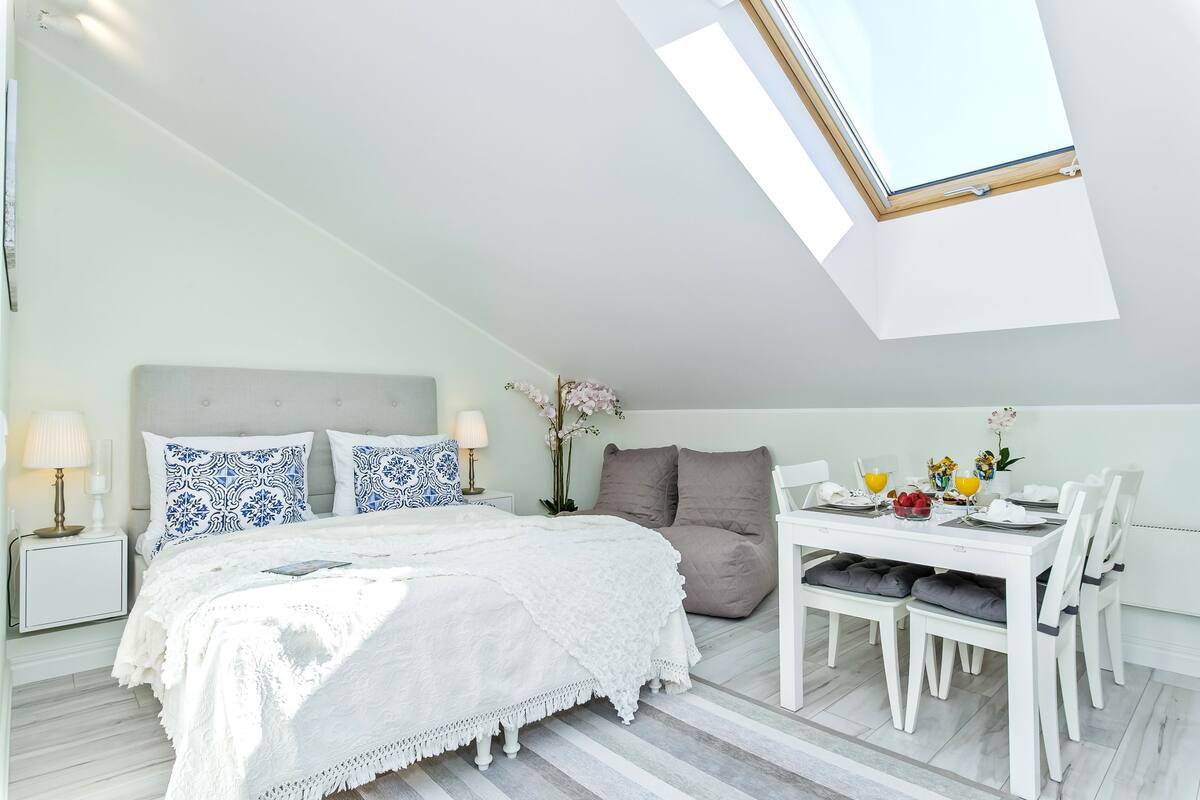 A bedroom with a skylight, a bed with patterned pillows, a bedside table with a lamp, and a dining area with chairs and a table set for two.